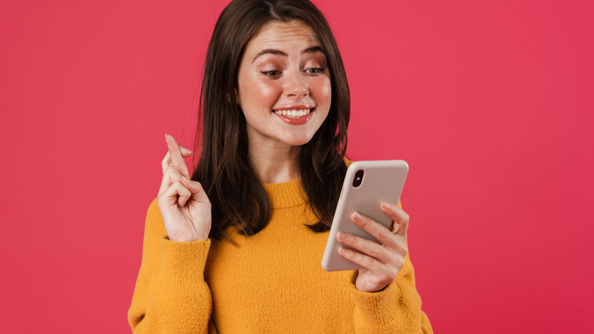 Imagem de fundo vermelho nela está uma mulher de cabelos médios e blusa amarela de frio segurando um celular com uma das mãos e na outra fazendo um sinal cruzando os dedos para algo dar certo.