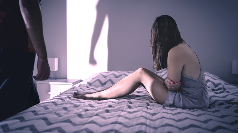 Mulher sentada em cama, acuada, e sombra de homem em pé na frente dela