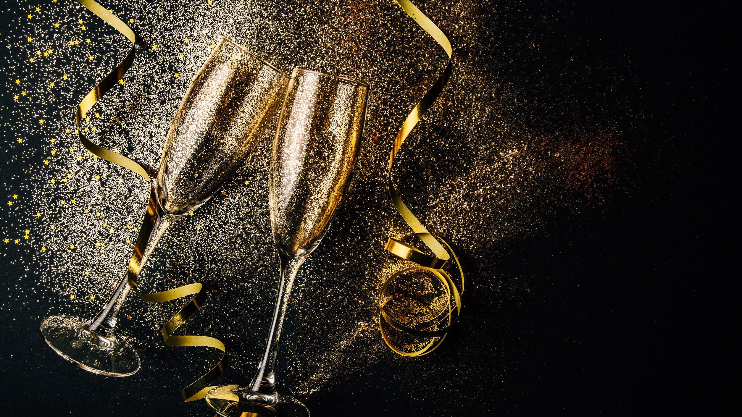 Taças de champanhe brindando em um fundo preto com glitter dourado espalhado e duas fitas douradas em espiral ao lado das taças.