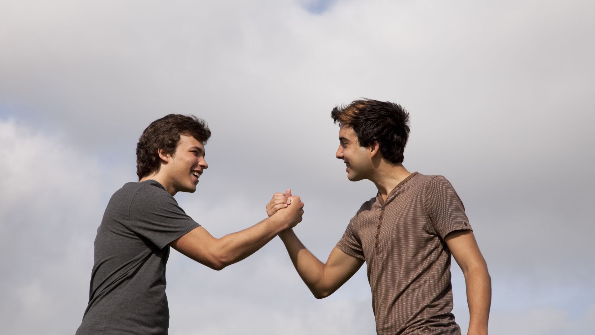 Imagem de dois homens se cumprimentando e sorrindo e o céu com nuvens atrás deles