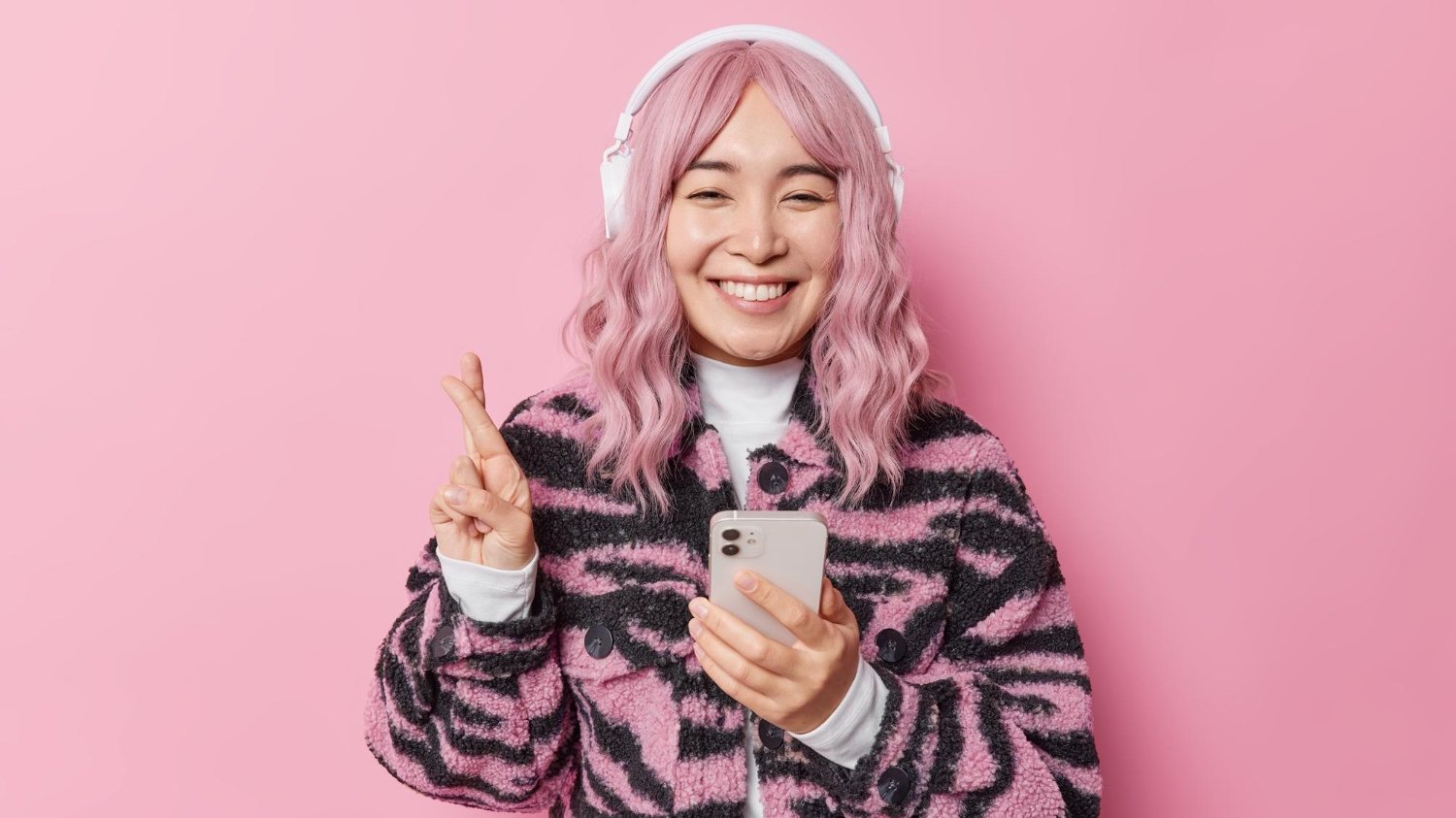 Mulher asiática com cabelo cor de rosa, fazendo sinal de com a mão e segurando o celular com a outra.