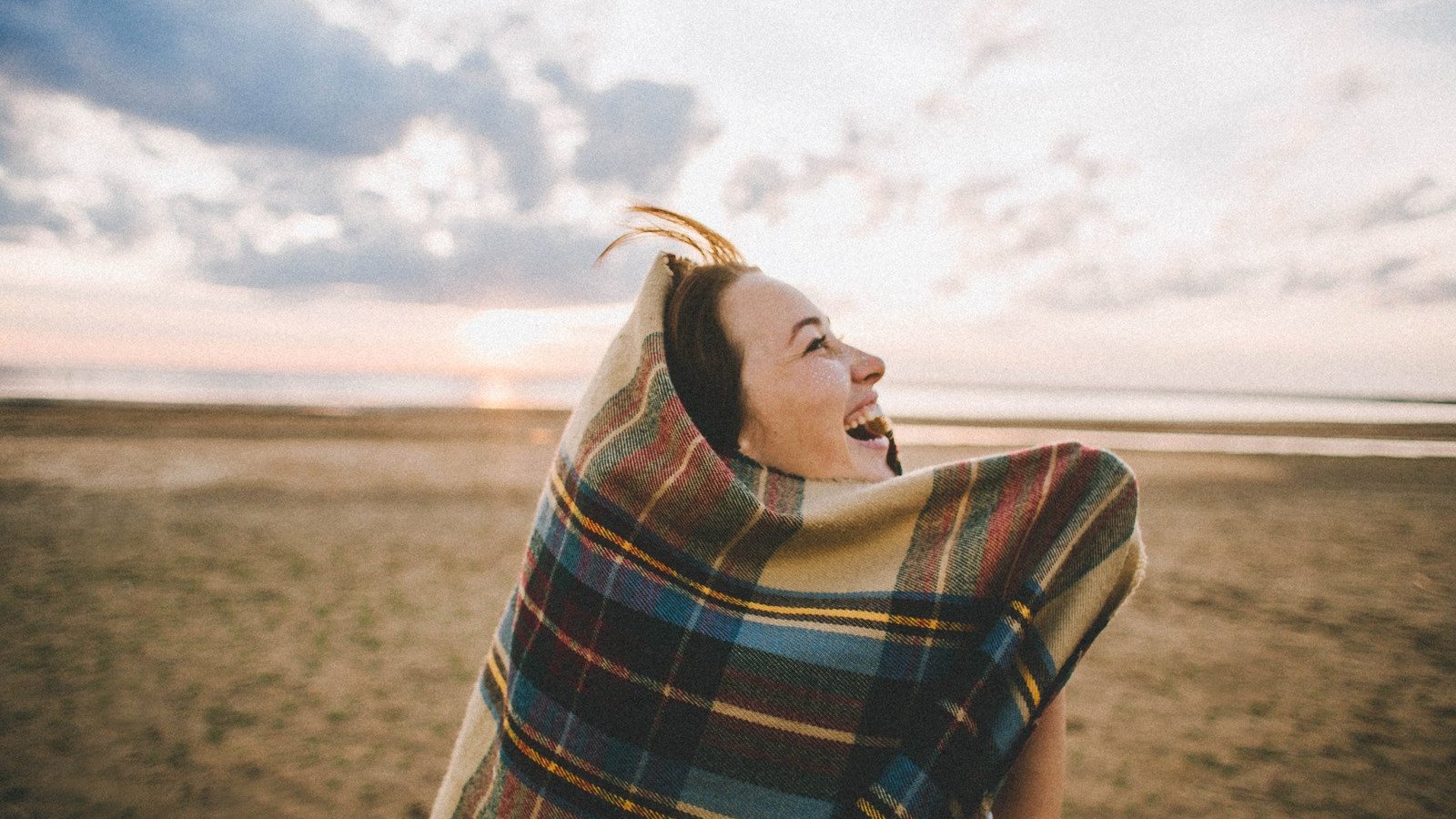 Valeria Ushakova / Canva PRO Mulher branca envolta de uma coberta na praia.