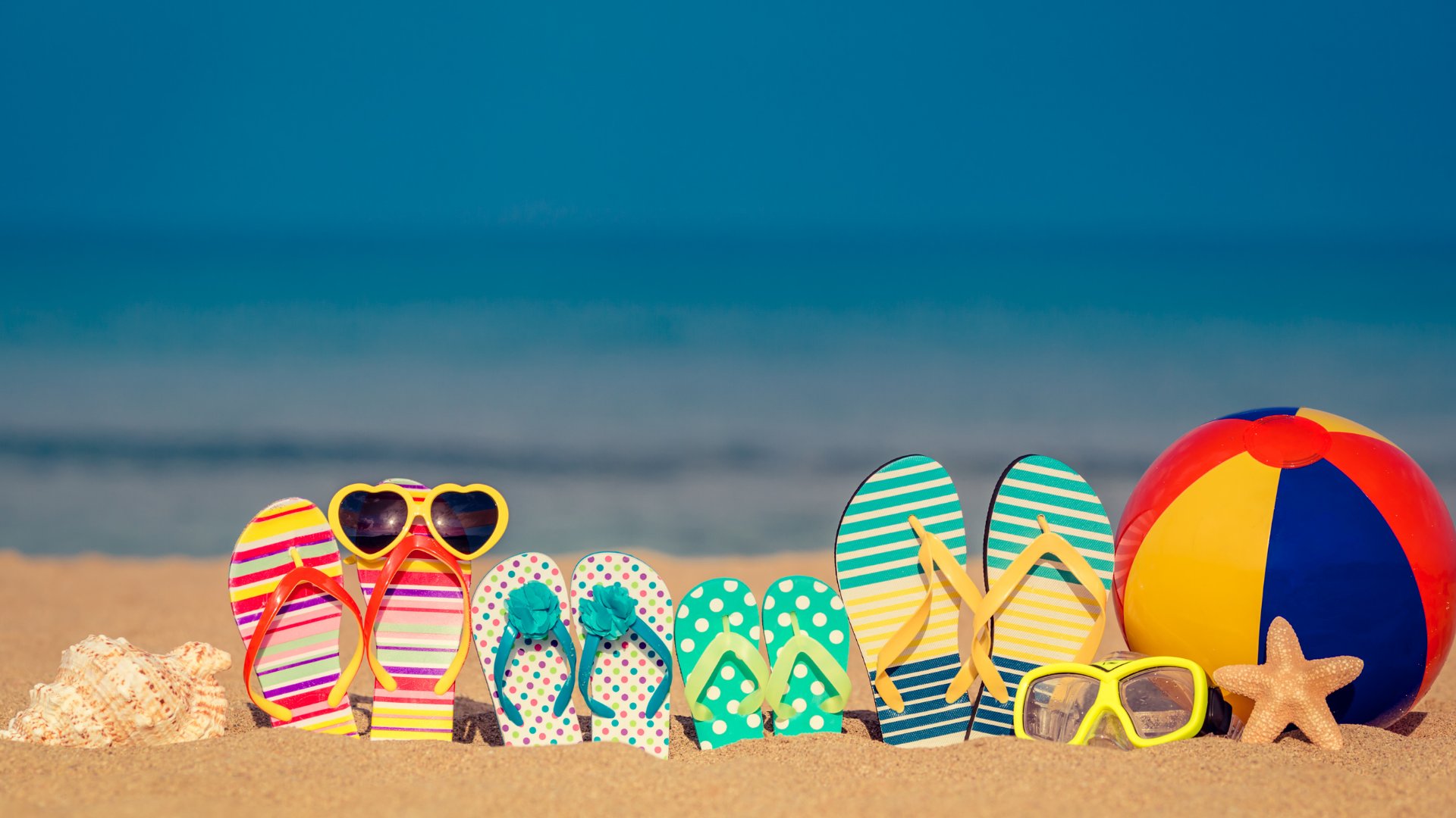 Chinelos de uma família enterrados na areia da paria junto de óculos de sol e bola de praia