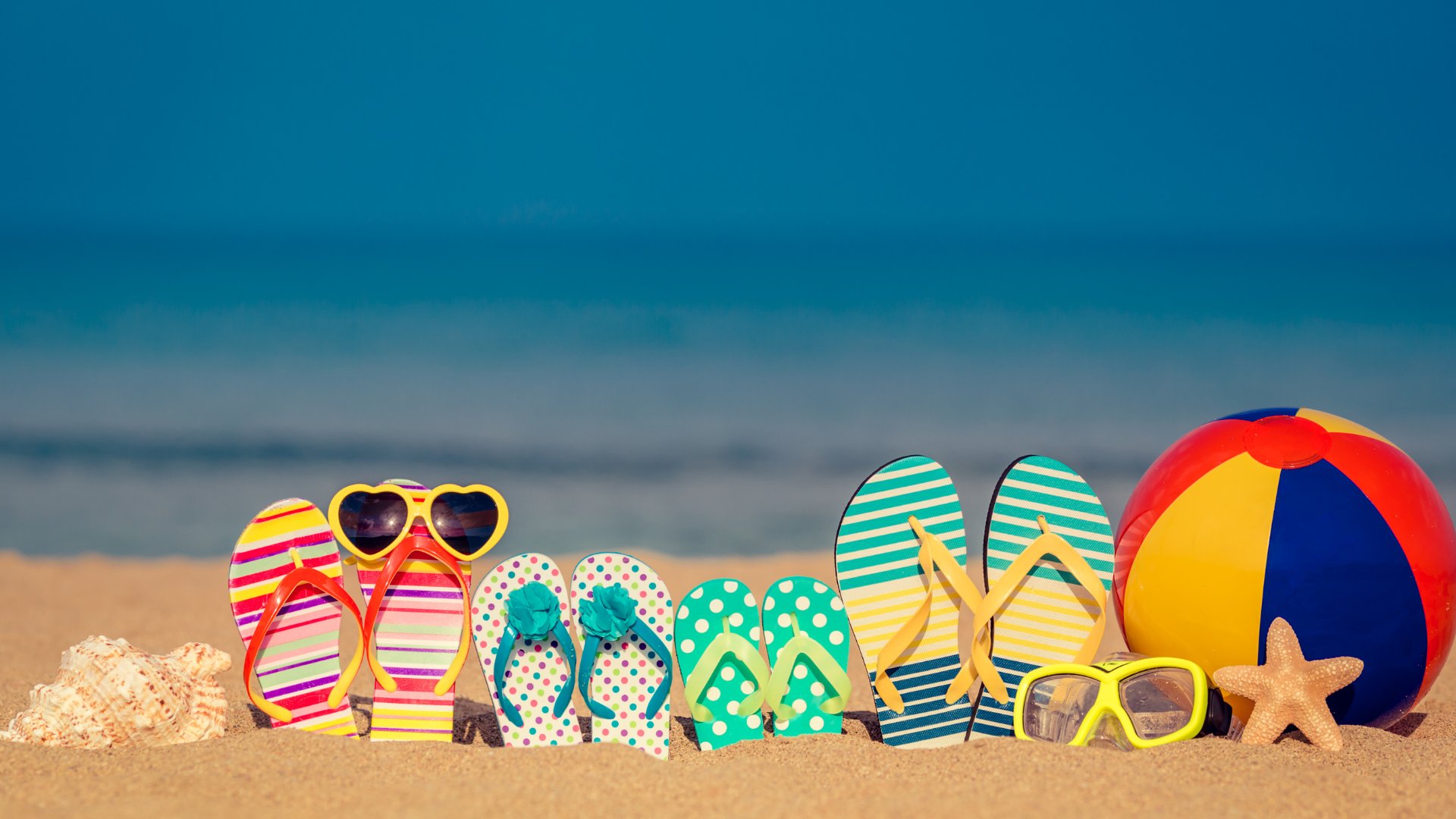 Yaruta / Getty Images Pro / Canva Chinelos de uma família enterrados na areia da paria junto de óculos de sol e bola de praia