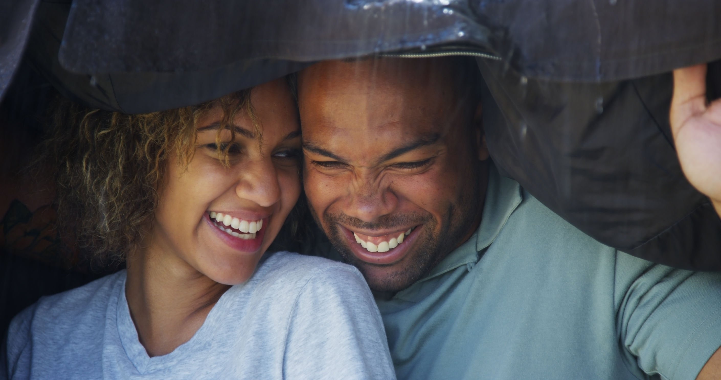Homem e mulher negros embaixo de casaco durante a chuva, com expressão sorridente.