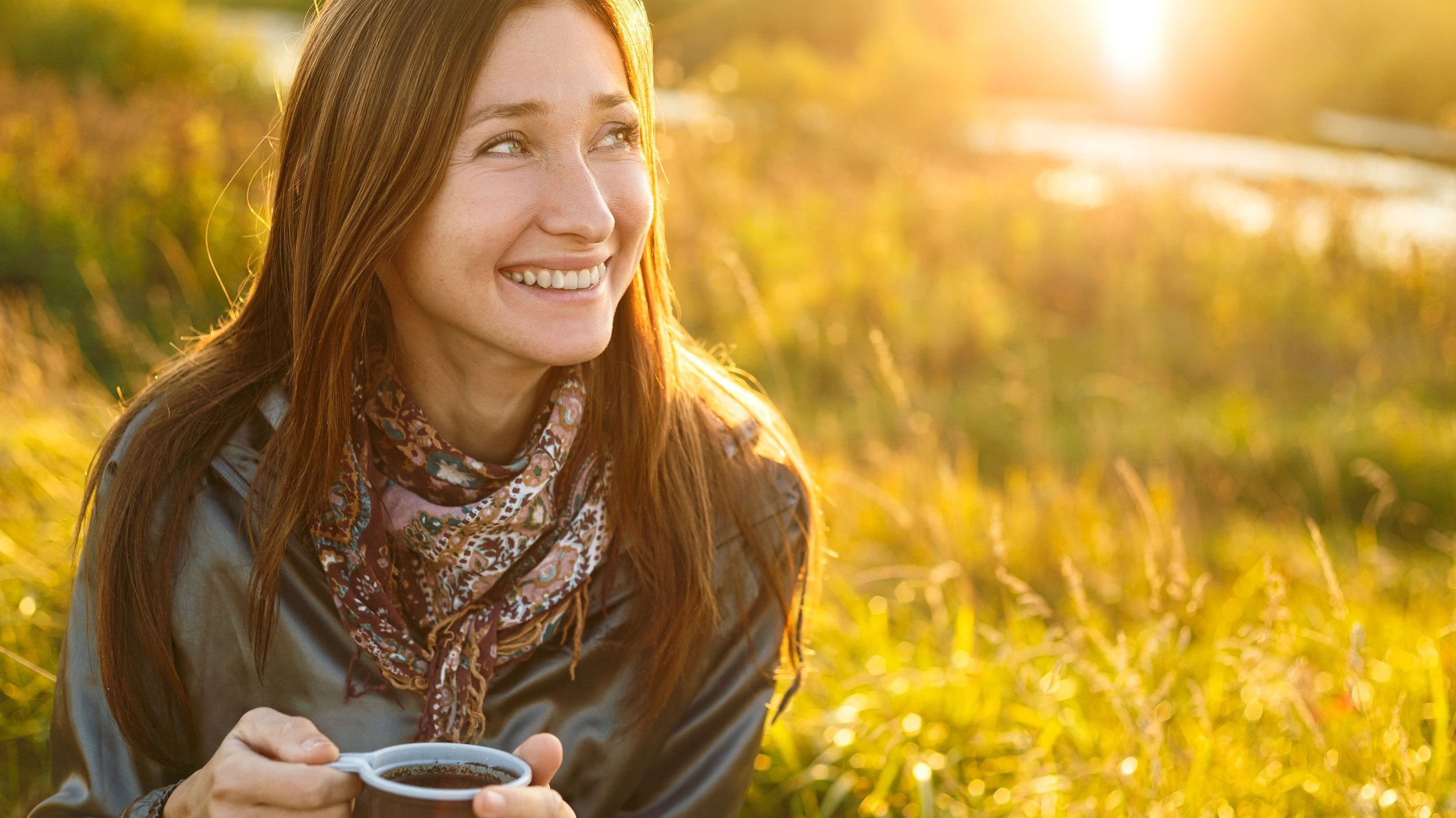 Mulher sentada em um gramado segurando uma xícara de café e sorrindo