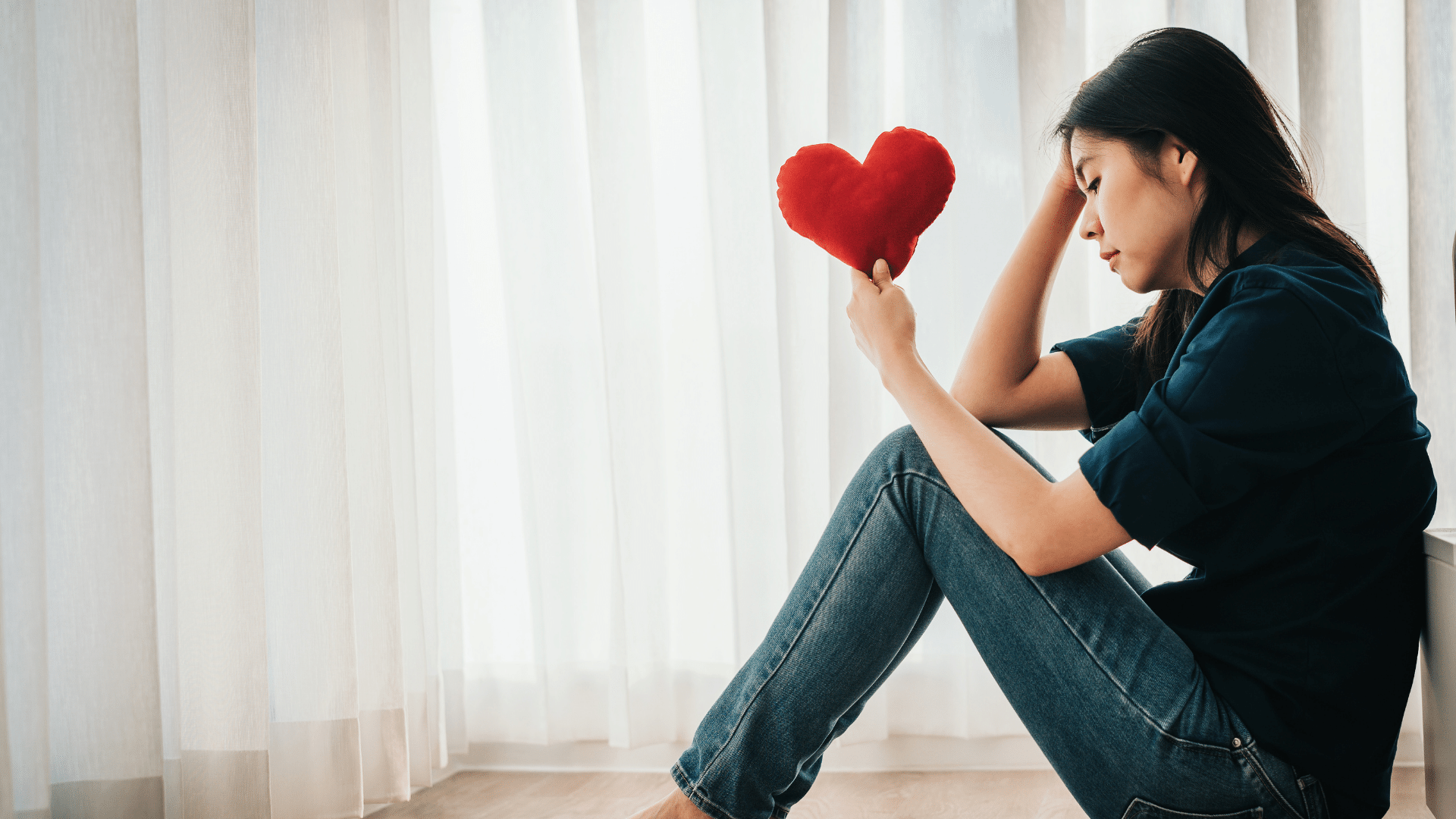 Mensagens Com Amor / Canva / interstid Foto de uma mulher sentada de cabeça baixa e semblante triste segurando um coração de pelúcia.