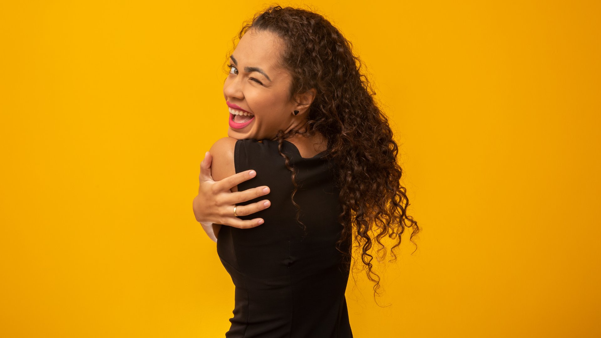 Cristiano Babini / Getty Images / Canva Mulher negra se abraçando enquanto sorri e pisca em um fundo amarelo