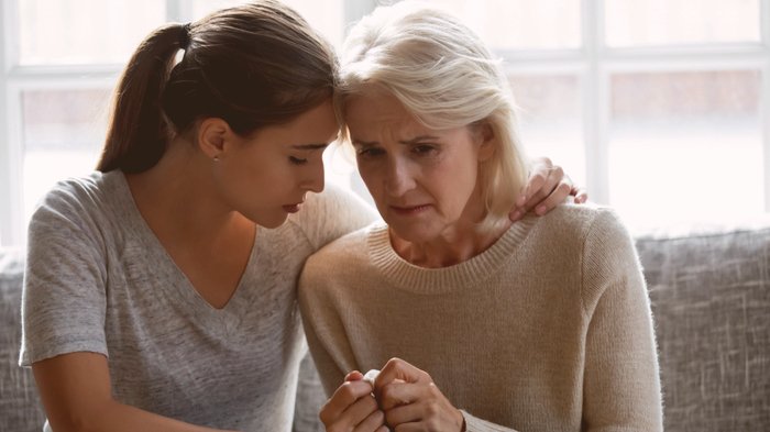 Jovem ao lado de idosa triste, que segura um lenço nas mãos. A jovem a abraça parcialmente e encosta sua cabeça na dela. Ambas estão sentadas em um sofá.