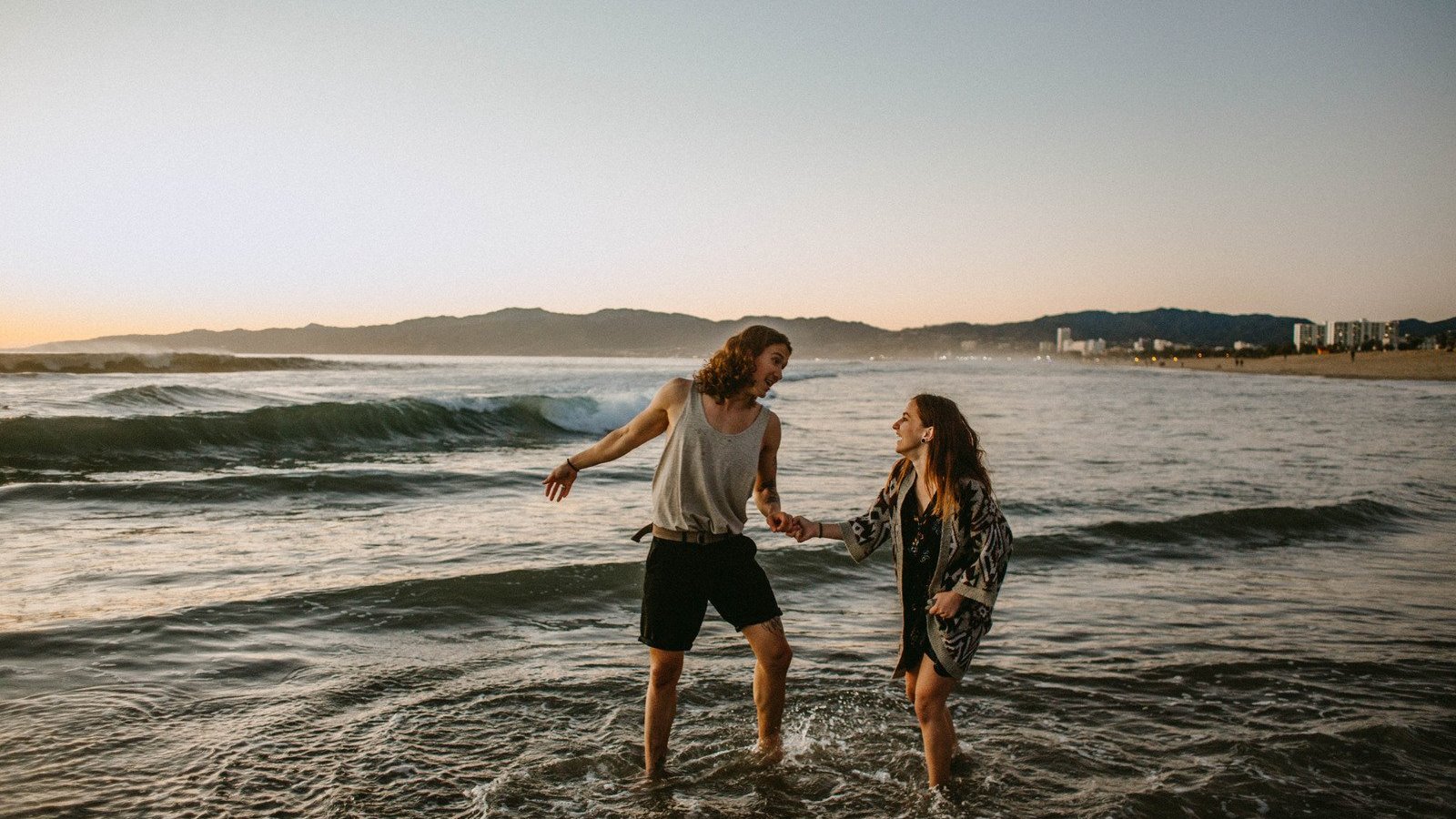 Homem e mulher brancos, de mãos dadas na beira do mar.