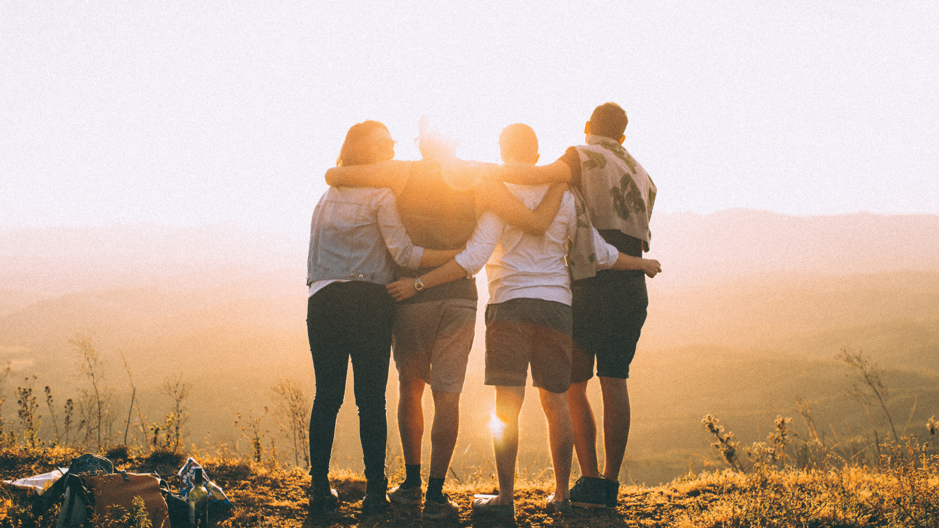 Imagem de quatro amigos de costas abraçados em uma paisagem de por do sol.