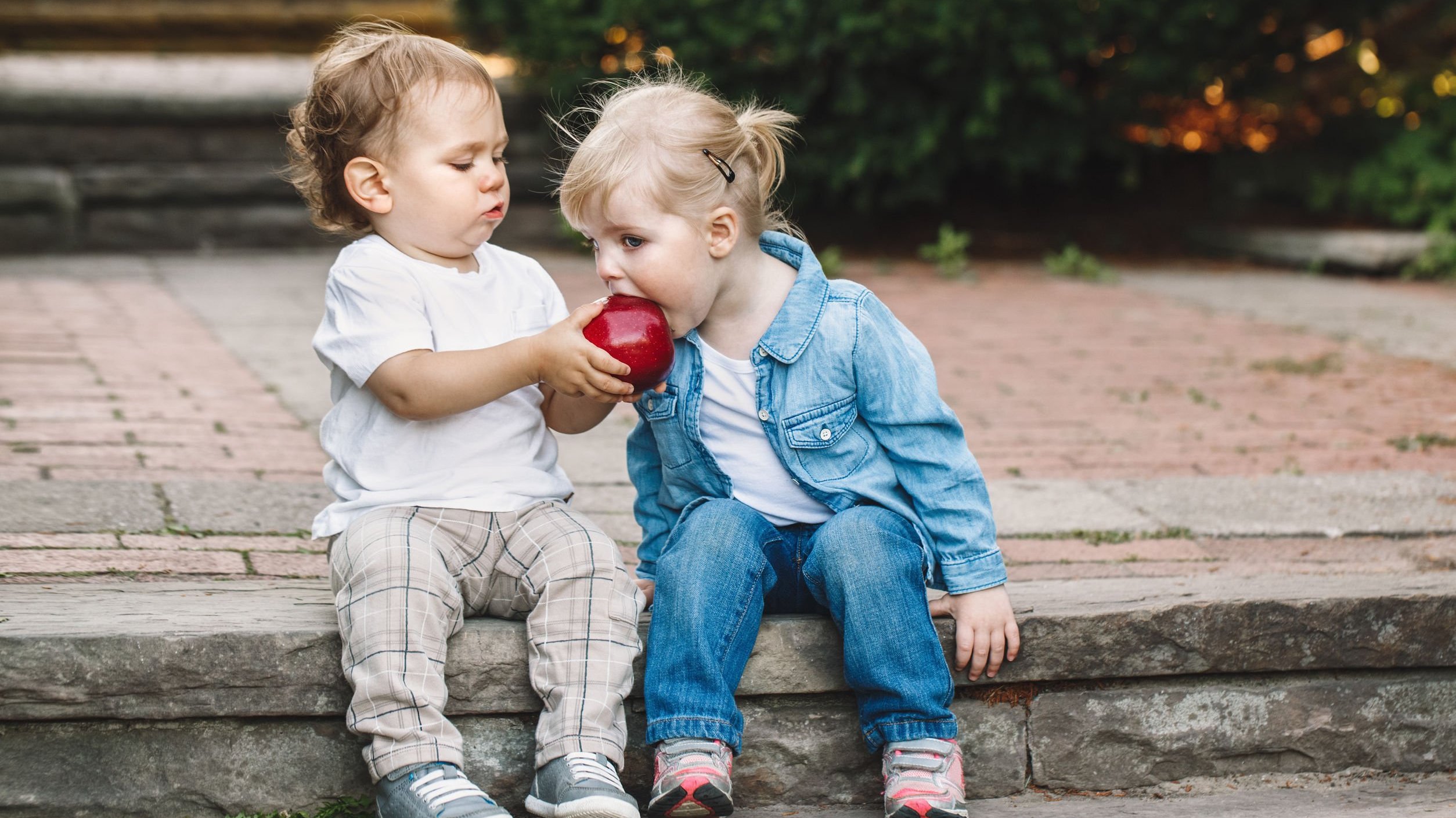 Menino e menina pequenos, sentados em uma sarjeta. O menino segura uma maçã, e a menina dá uma mordida nela.