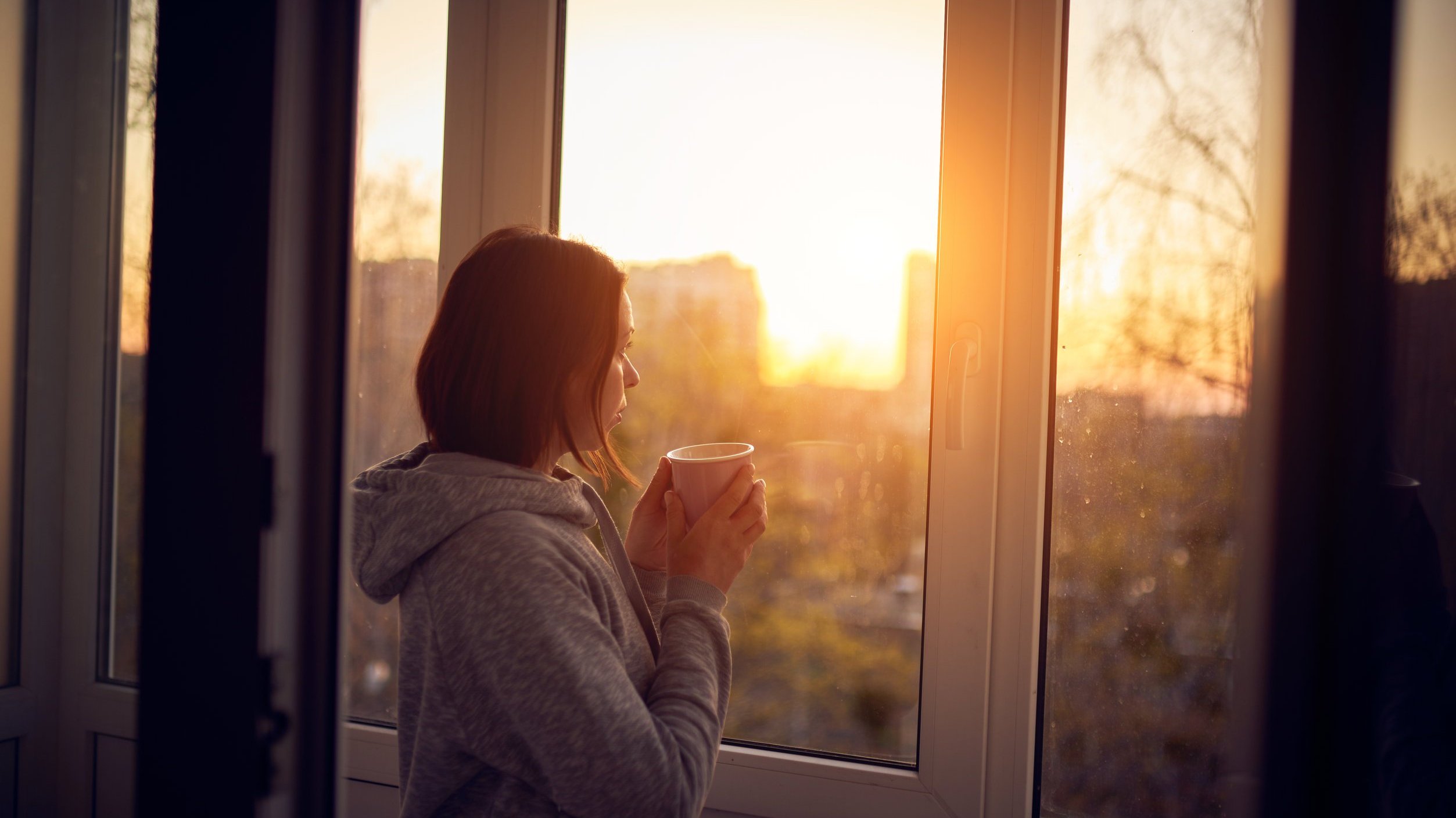 Mulher segurando uma xícara de café de frente para uma janela
