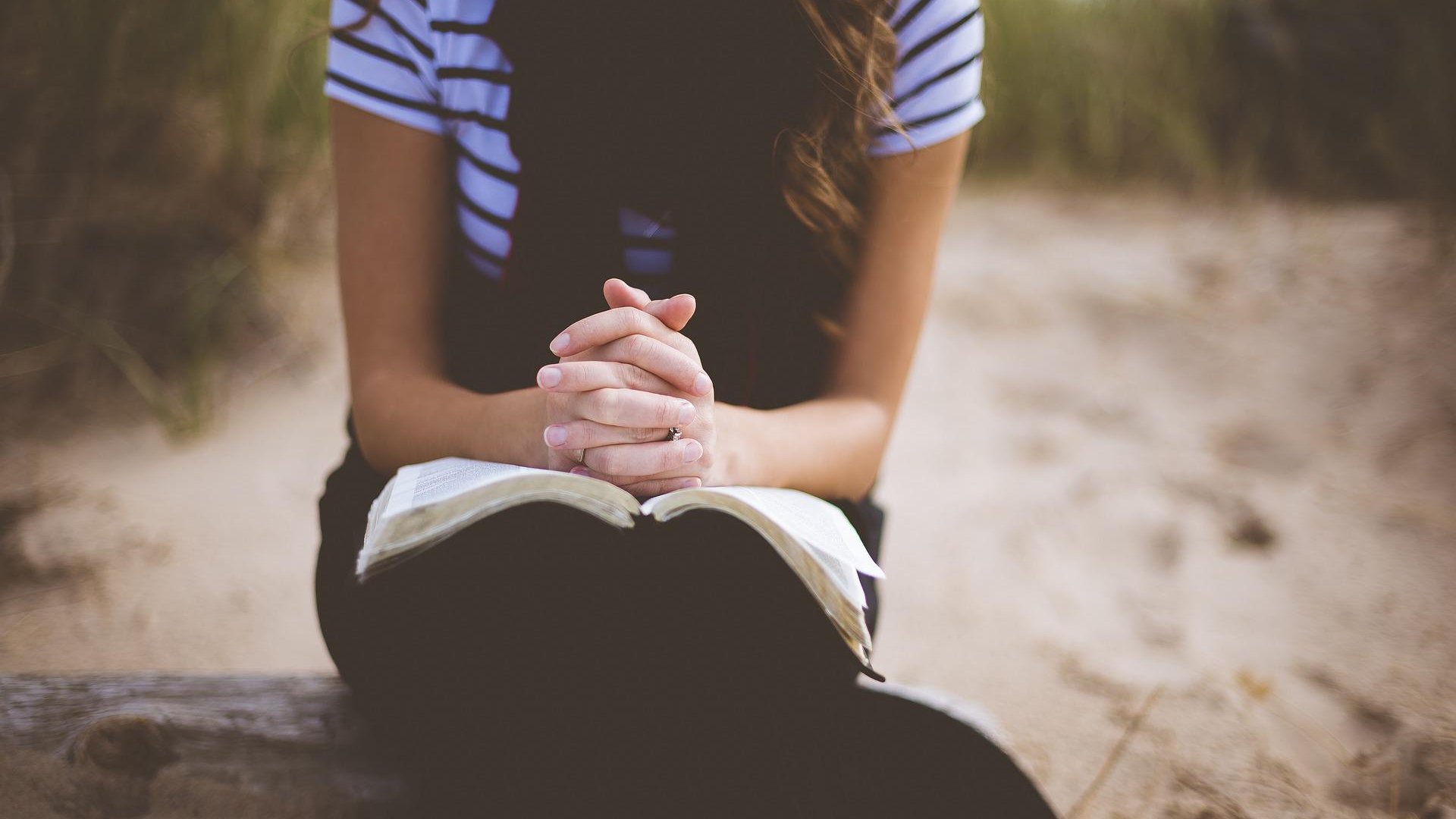 Na praia, uma moça está rezando com uma Bíblia apoiada em seu colo.