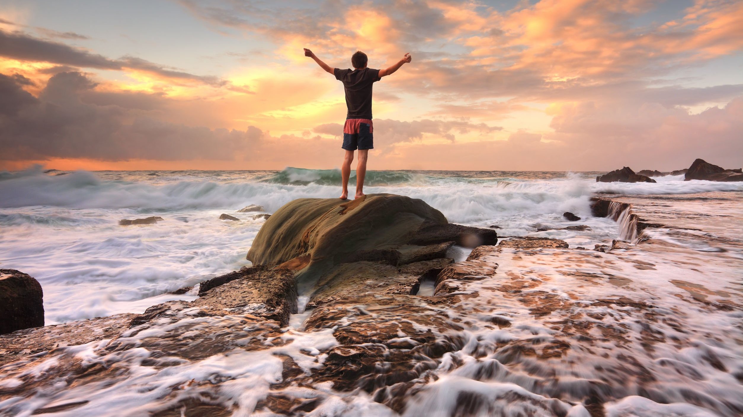 Homem de pé em uma pedra, na praia, de braços abertos.