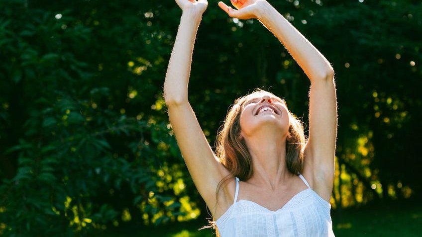 Mulher com braços para cima sorrindo com fundo de folhar verdes sob o sol