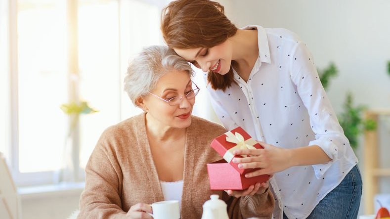 Senhora sentada em mesa com sobrinha em pé lhe mostrando presente