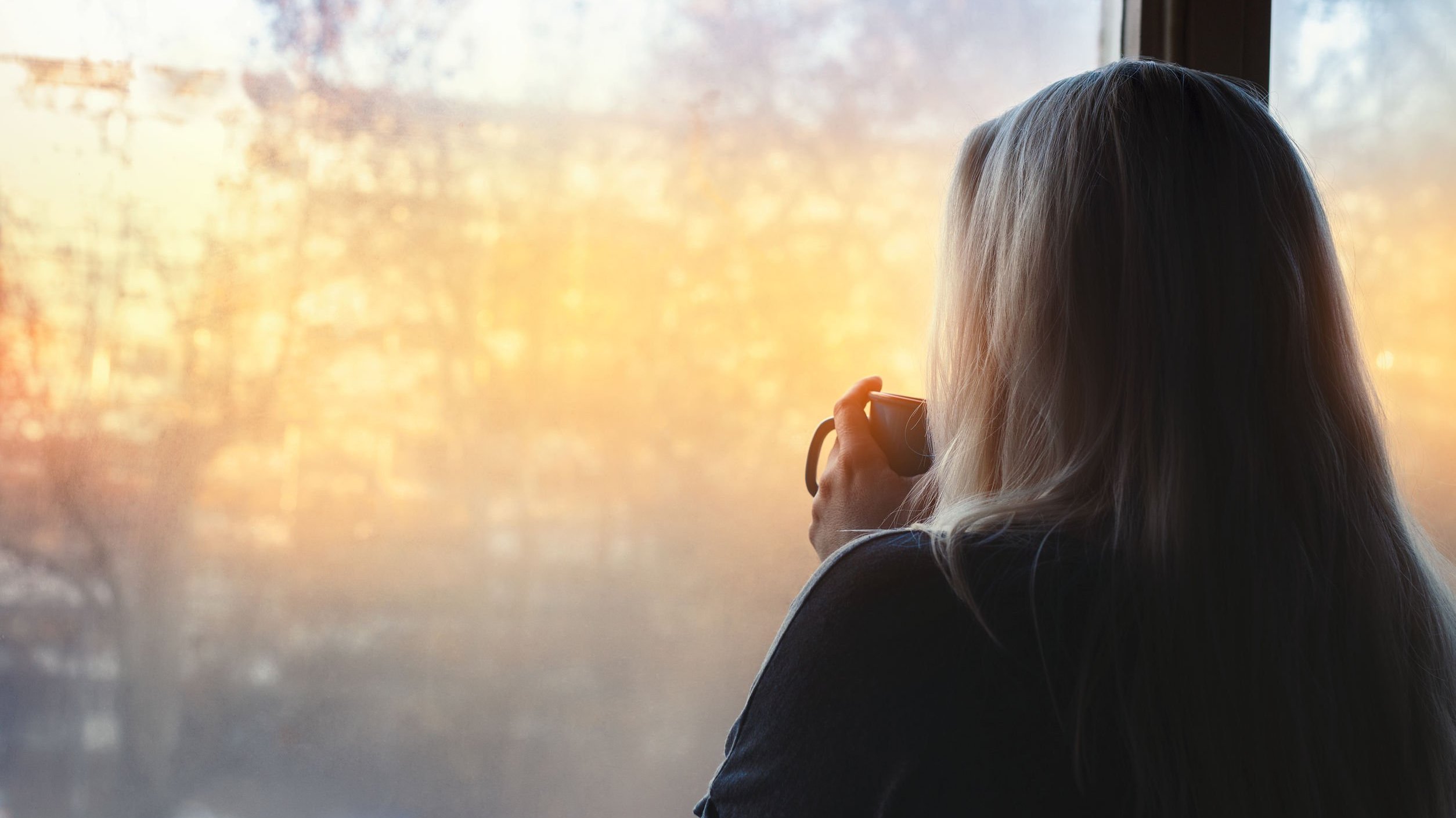Mulher de costas para a câmera, olhando para uma janela que mostra árvores e a luz amarela do sol, enquanto segura uma caneca.