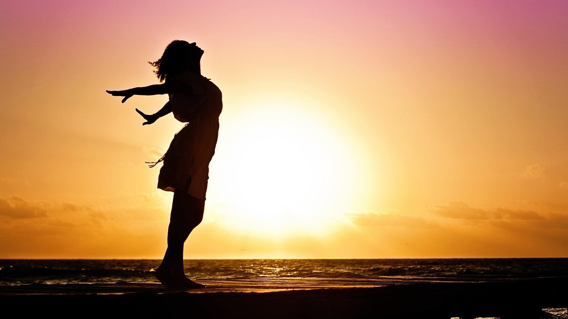 Mulher está de braços abertos olhando para o céu. O sol está se pondo atrás dela.