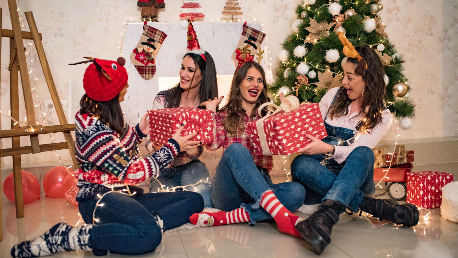 Imagem com elementos decoarativos de natal e 4 amigas sentadas no chão, usando tiaras temáticas e trocando presentes.