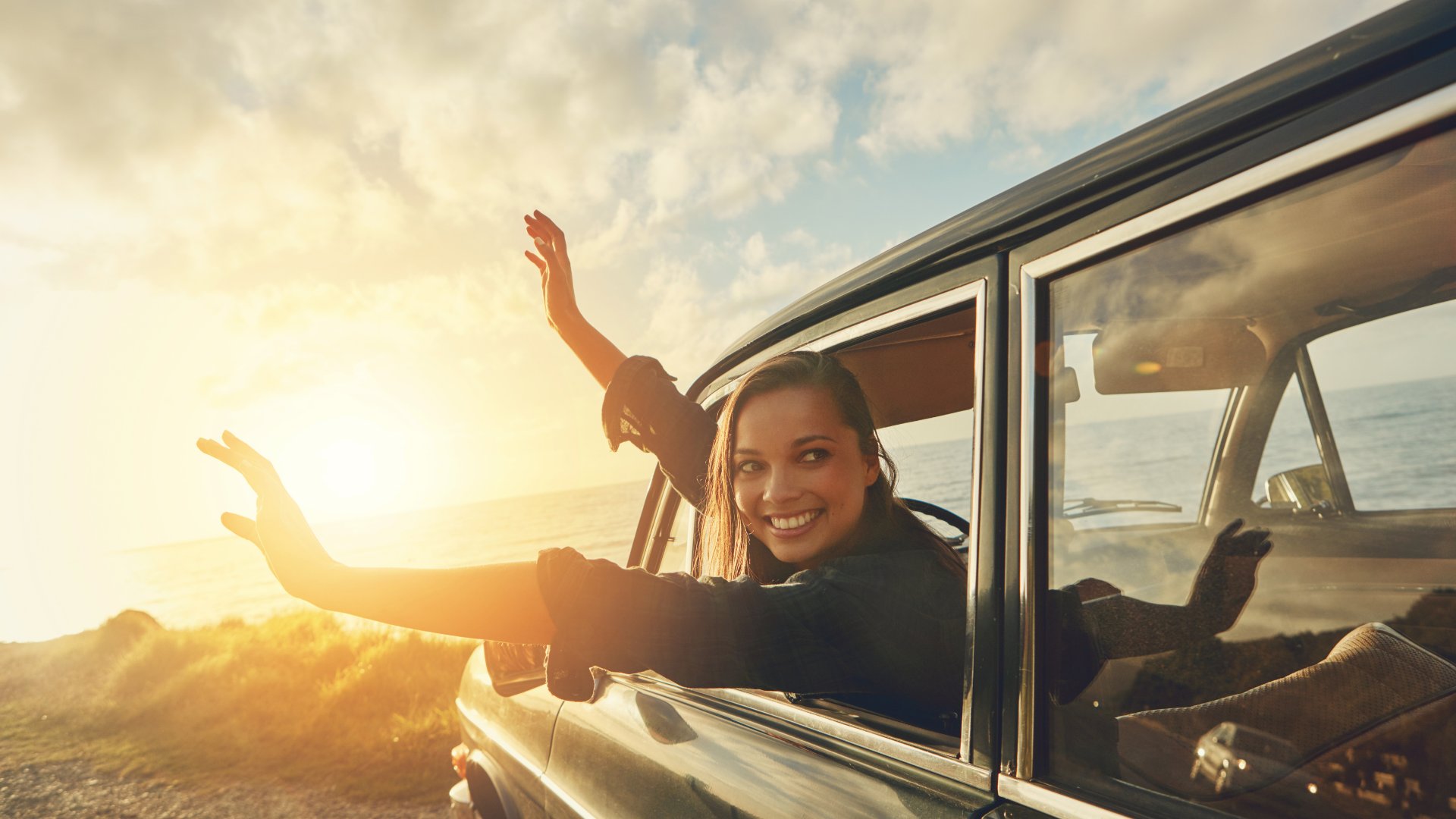 Mulher sorrindo dentro de um carro com as mãos para fora da janela, observando o pôr do sol