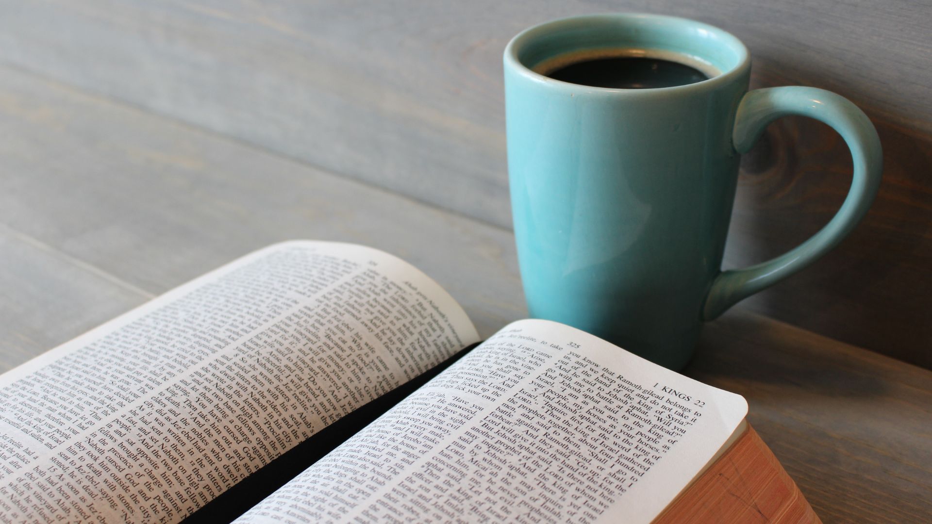 Imagem de uma bíblia aberta sobre uma mesa de madeira clara e à frente dela, uma caneca na cor azul cheia de café.