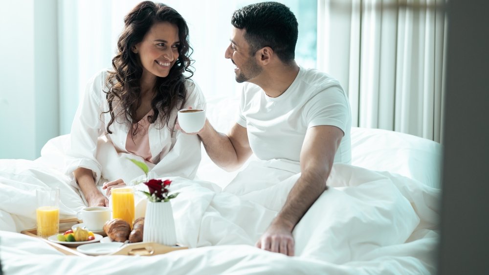 Casal sentado e tomando café da manhã na cama.