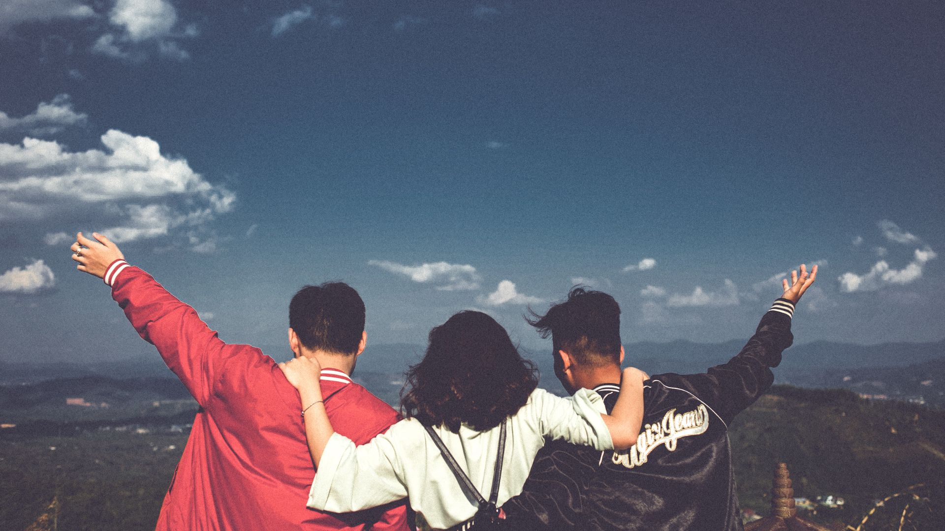 Imagem de três amigos no topo de uma montanha, com o céu azulado a frente e abraçados