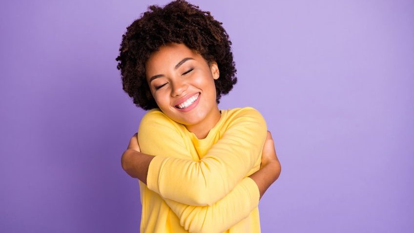 Foto de uma jovem abraçando-se e sorrindo com o fundo de cor roxa