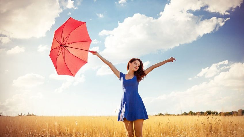 Mulher com vestido azul e braços abertos segurando guarda-chuva vermelho em campo e céu ao fundo