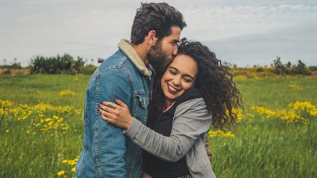 Casal se abraçando em meio a um campo esverdeado. A mulher está sorridente.