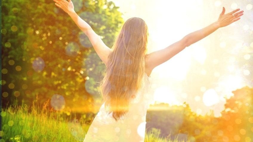 Mulher com cabelos compridos e usando um vestido, de costas para a câmera com os braços erguidos para o alto, em um campo gramado com árvores. A luz do sol atinge a mulher e a lente da câmera.