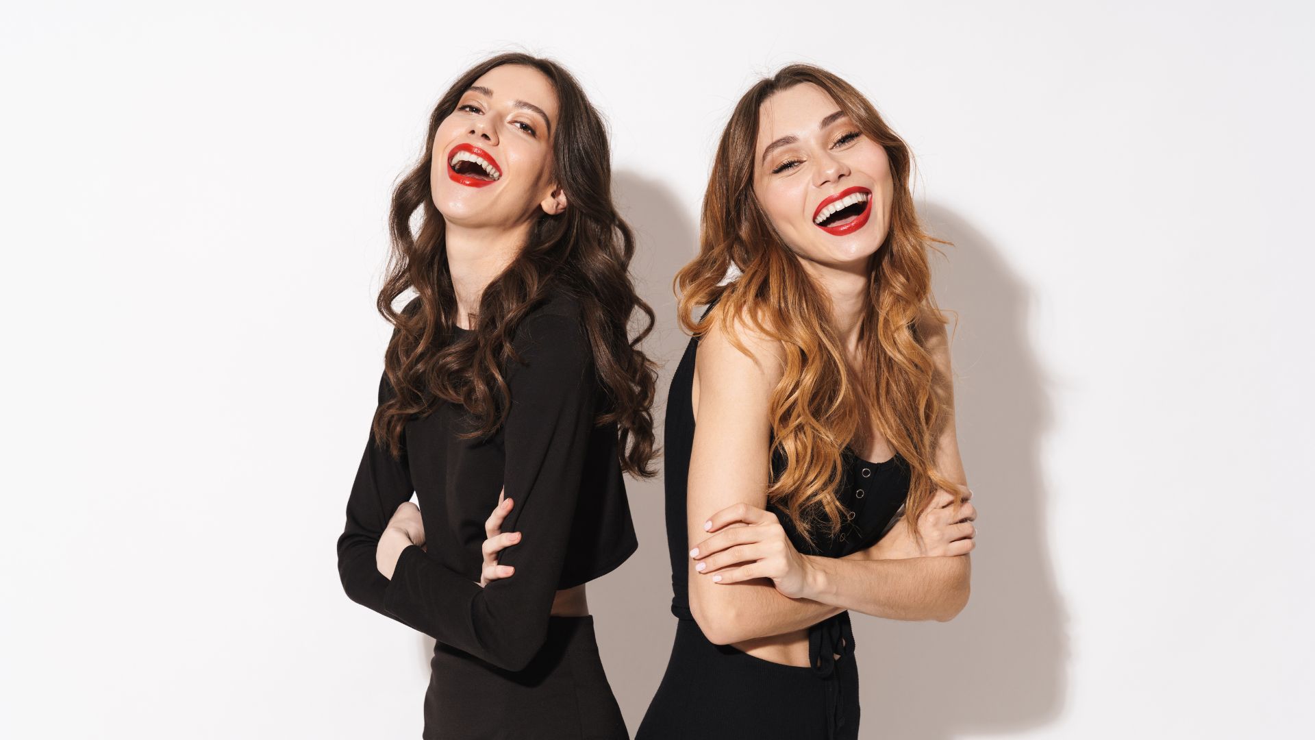 Imagem de duas mulheres de braços cruzados viradas de costas uma para outra, sorrindo.