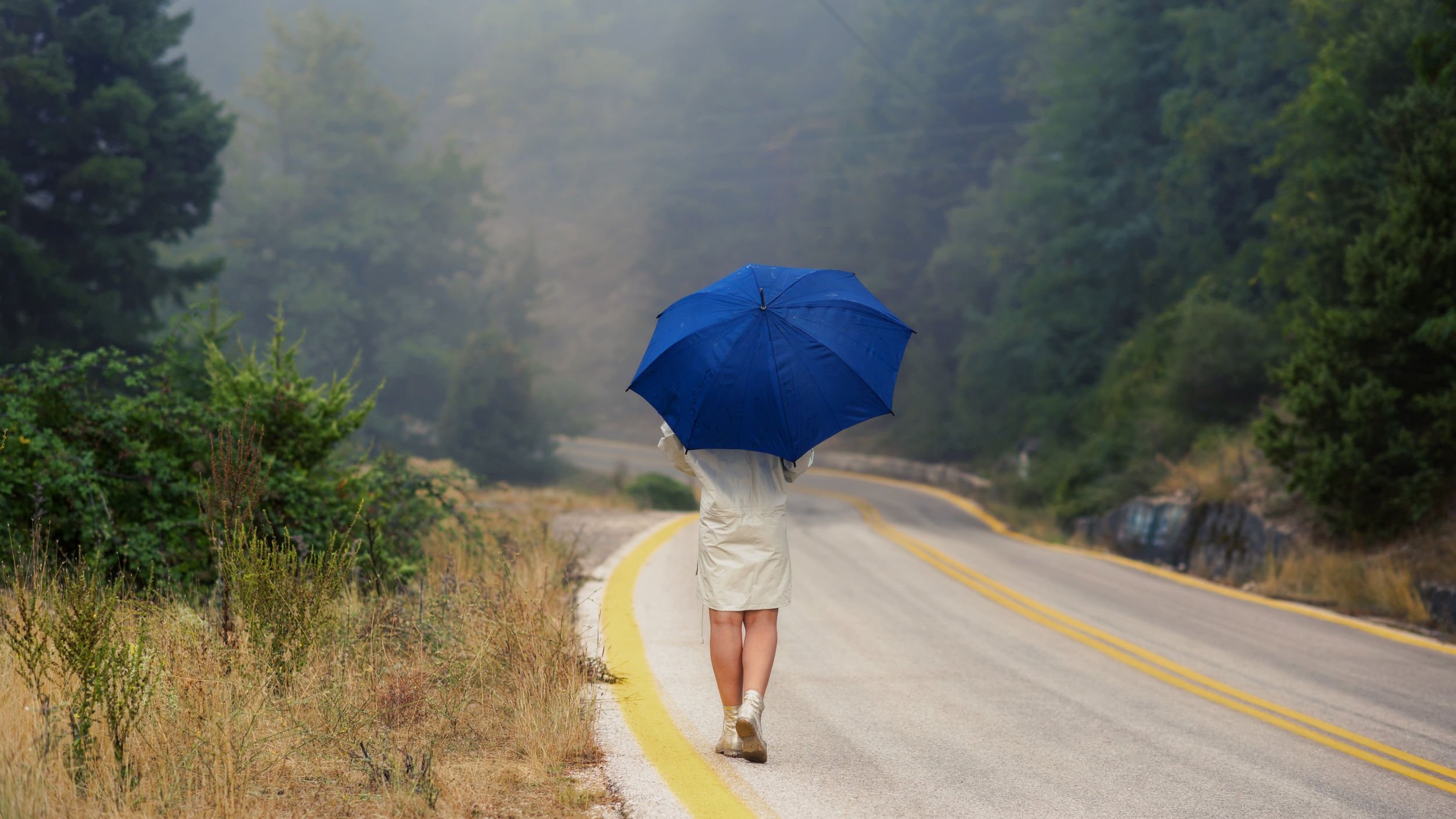 Mulher com capa de chuva e segurando um guarda-chuva, andando em uma rodovia deserta.