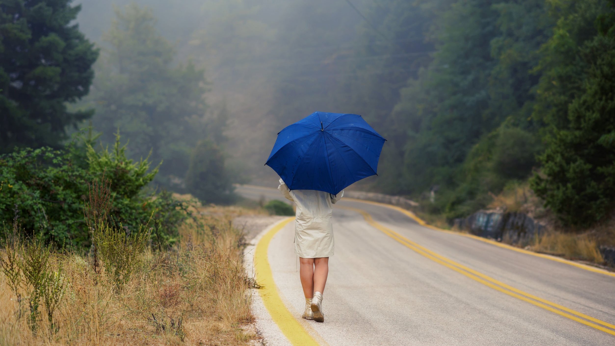 123rf/flowertiare Mulher com capa de chuva e segurando um guarda-chuva, andando em uma rodovia deserta.