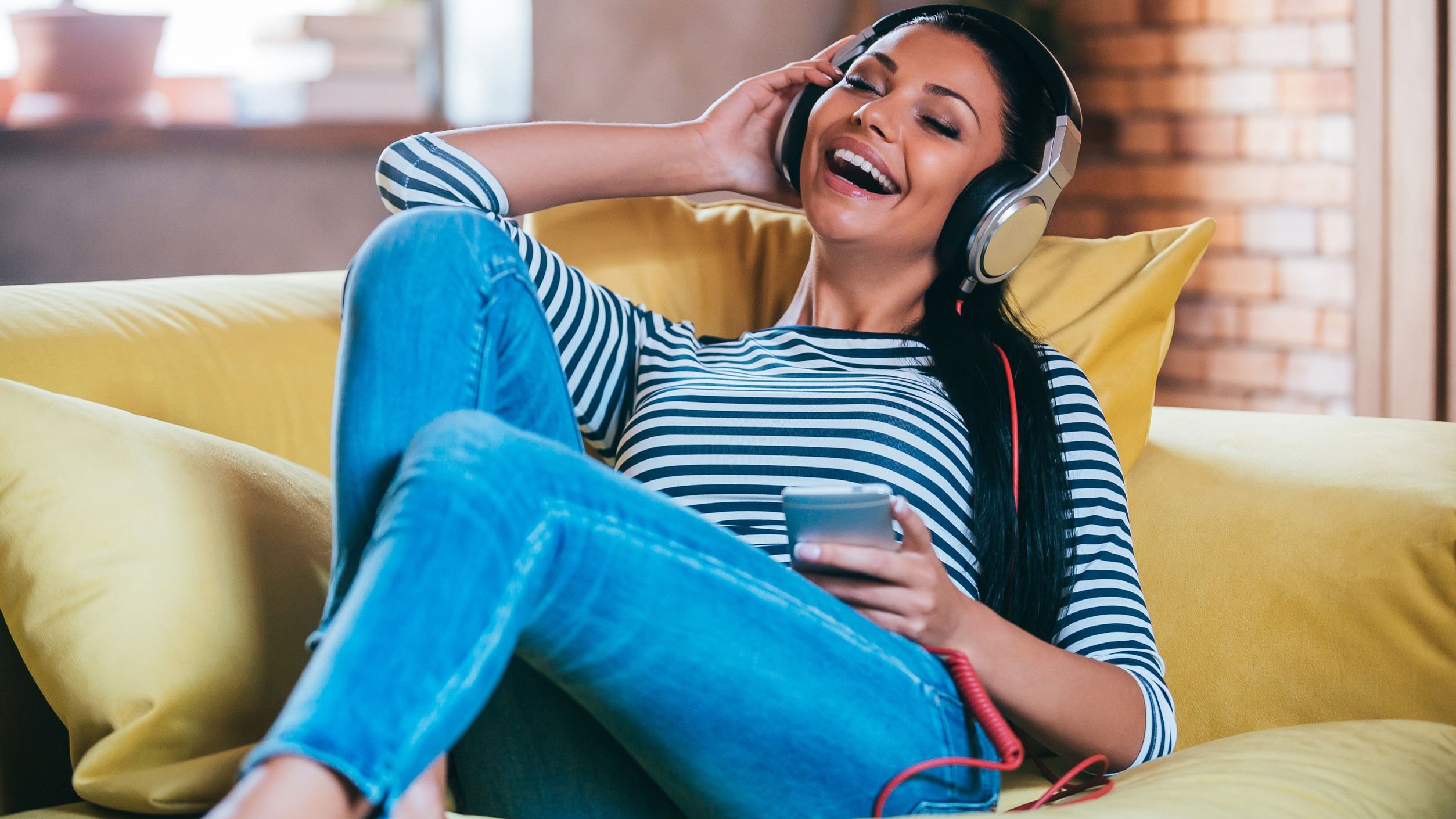 Mulher branca deitada num sofá amarelo com fones de ouvido.