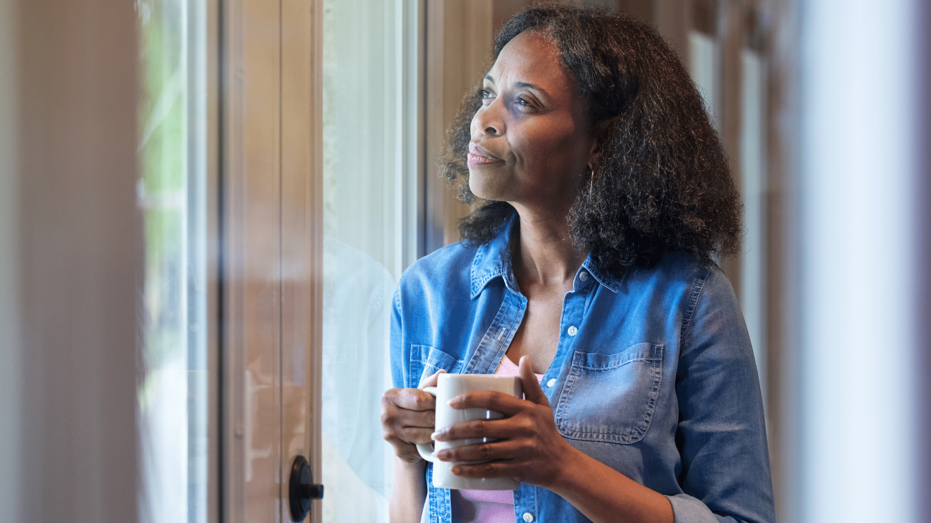 Mulher olhando pela janela e refletindo com uma caneca de café na mão.