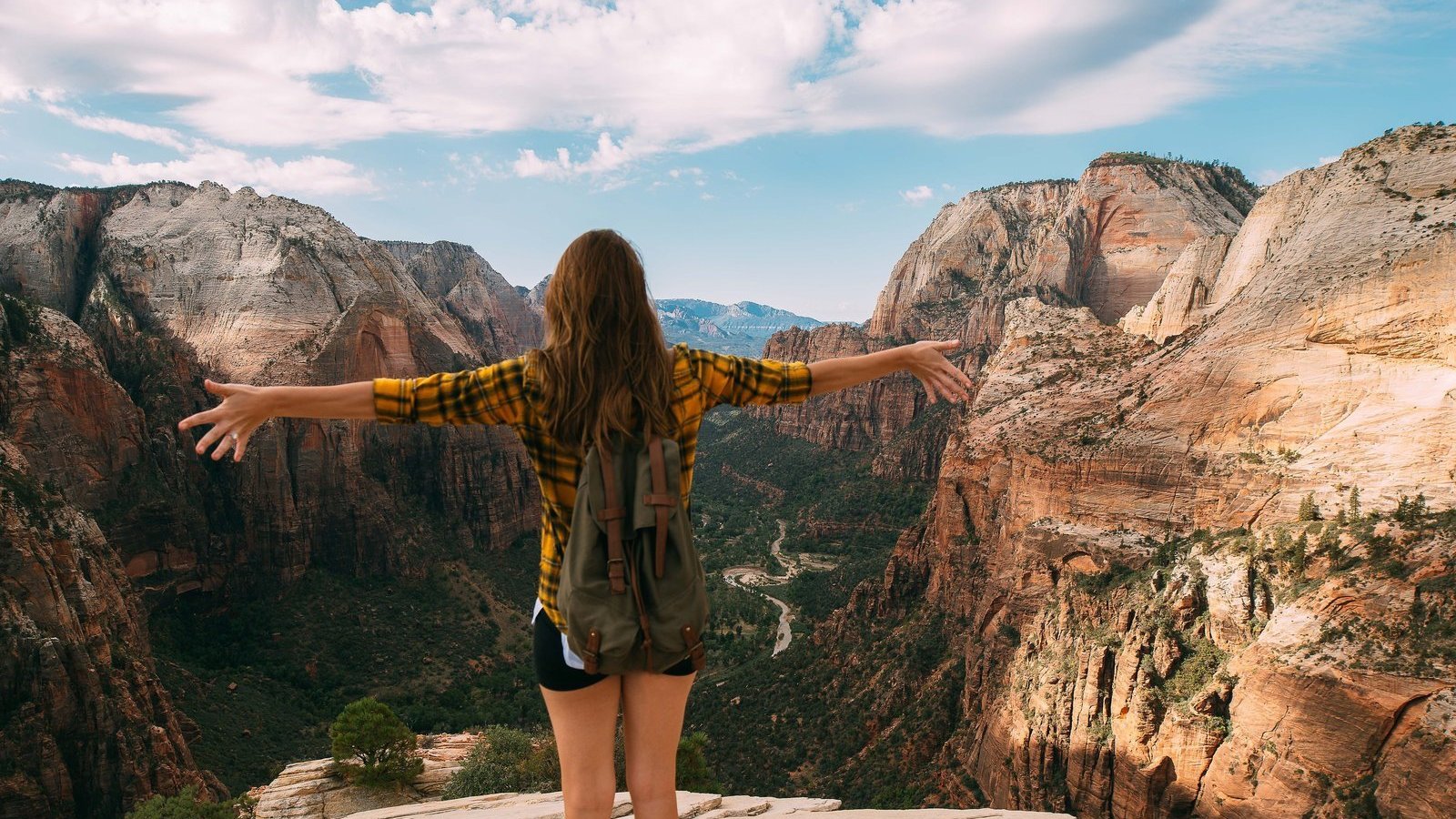 Menina de costas com camiseta xadrez amarela, mochila nas costas e shorts de braços abertos olhando para as montanhas.