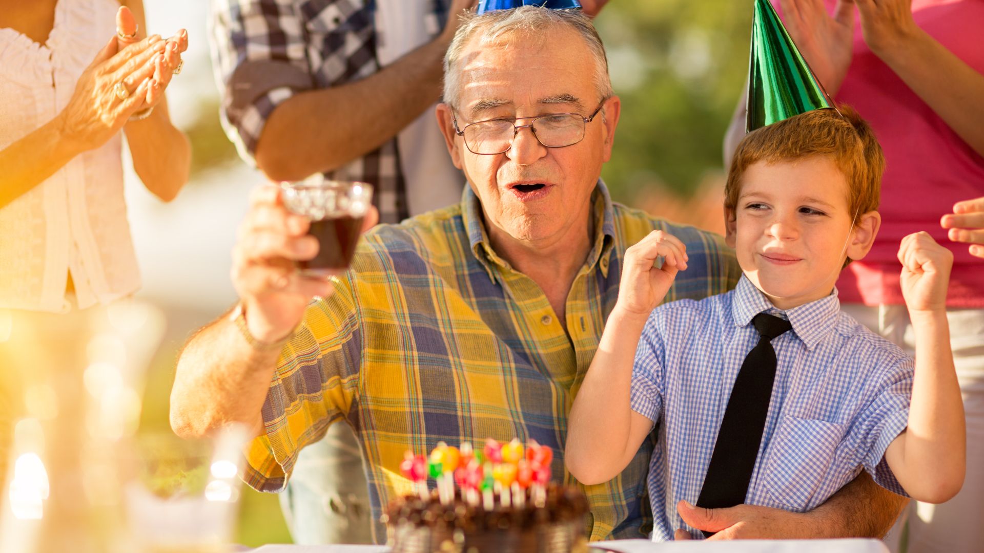 Imagem de um avô segurando seu neto no colo e eles estão com chapéus de aniversário na frente do bolo, eu uma festa