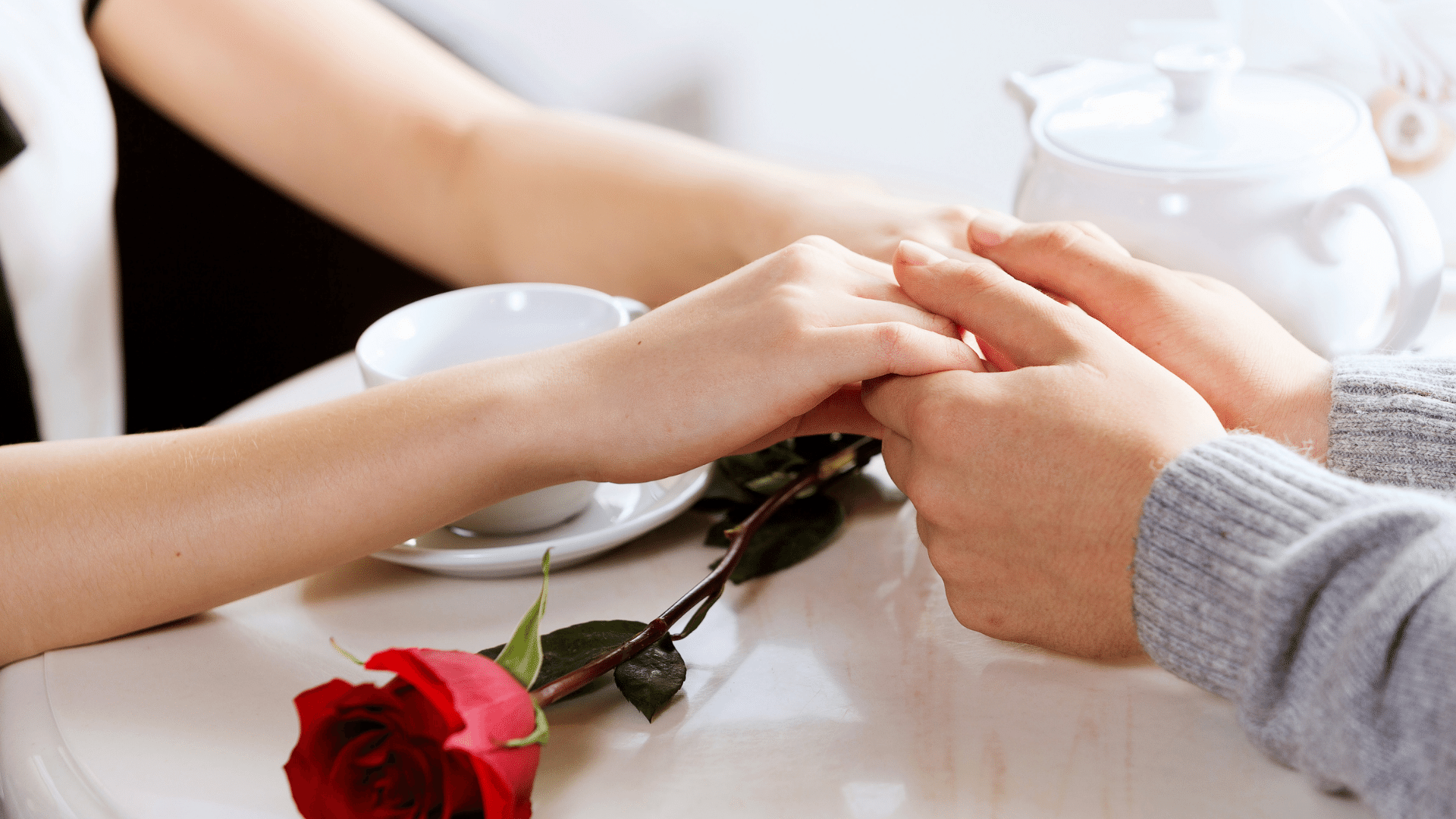Casal de mãos dadas na mesa de café da manhã e uma rosa vermelha em cima da mesa. Na foto só aparecem as mãos.