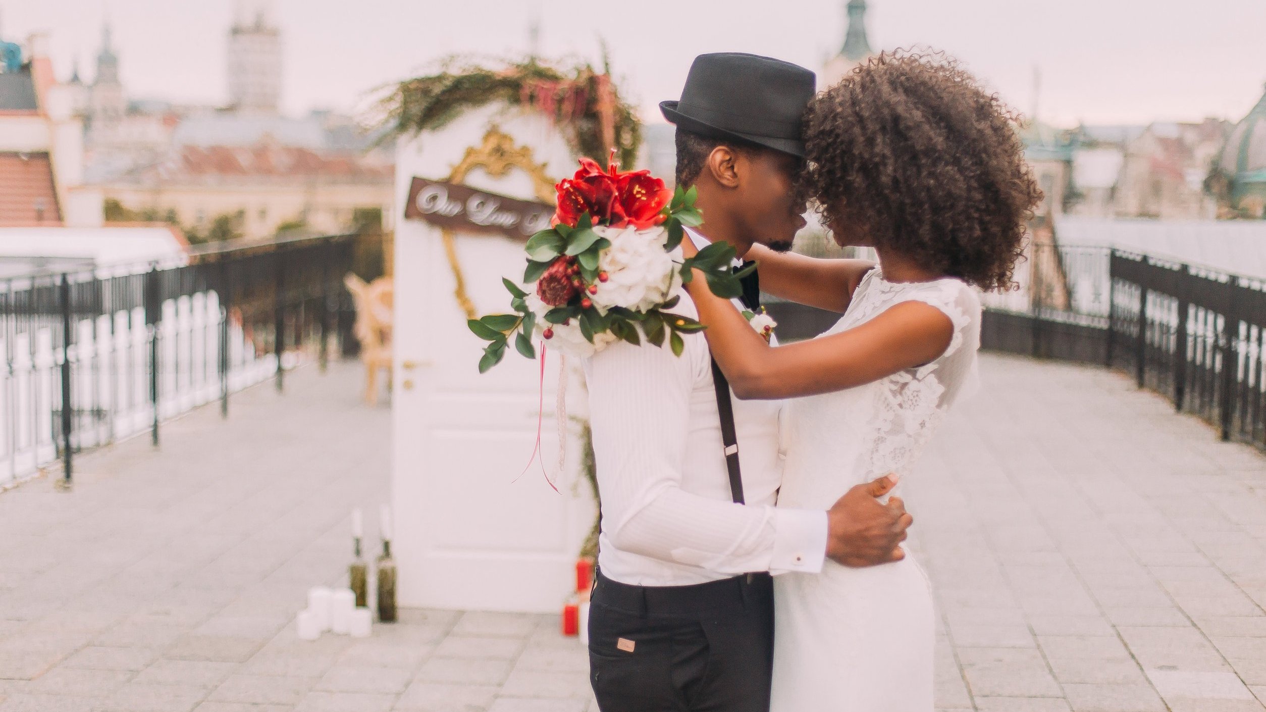 Um homem segurando na cintura de uma mulher que segura em volta da sua nuca, os dois estão com as testas coladas e vestidos de branco