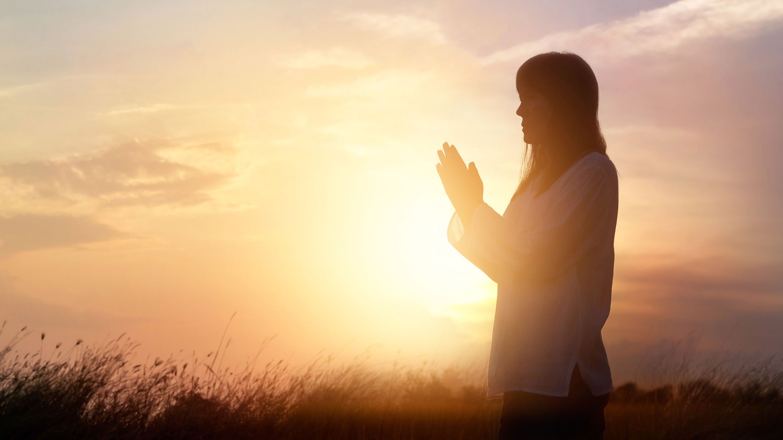 Silhueta de mulher orando, de perfil, em um campo, sob o pôr do sol.