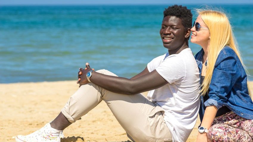 Casal sentado na areia da praia sorrindo no sol