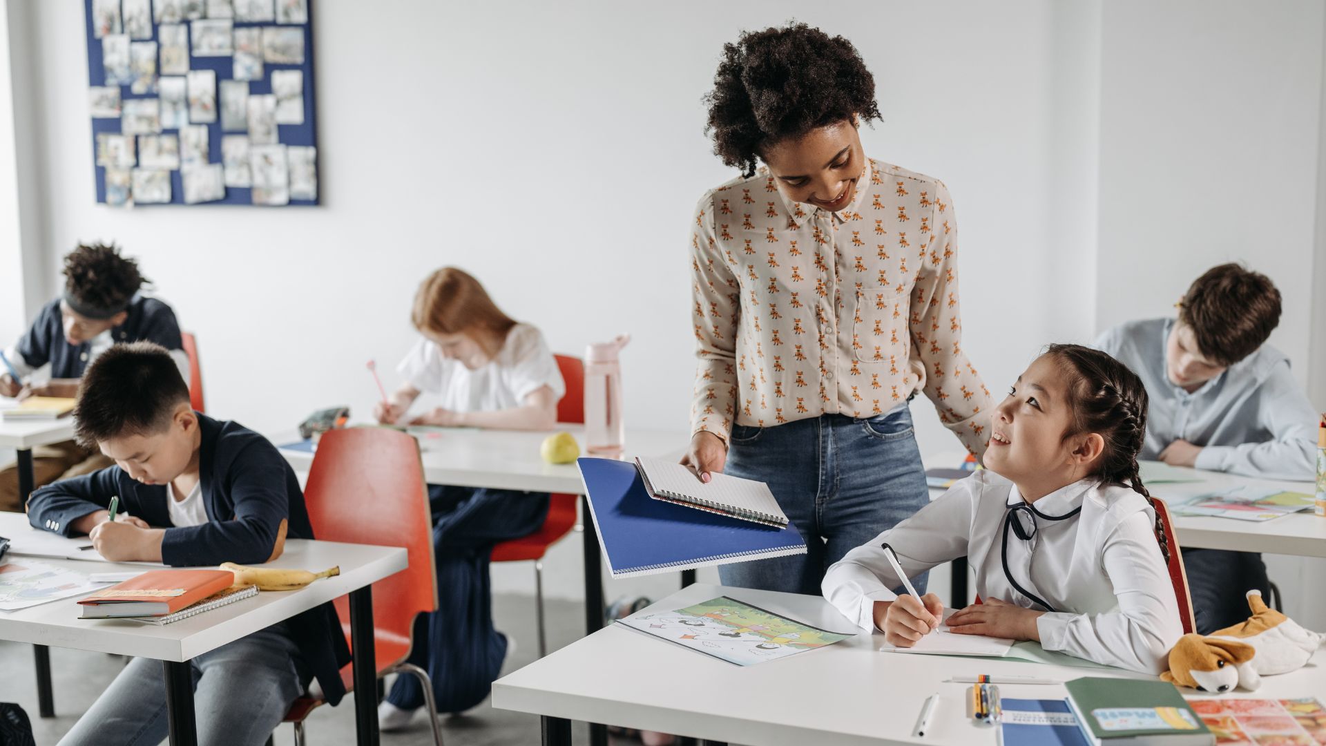 Imagem de uma professora dando aula na sala e ajudando uma criança