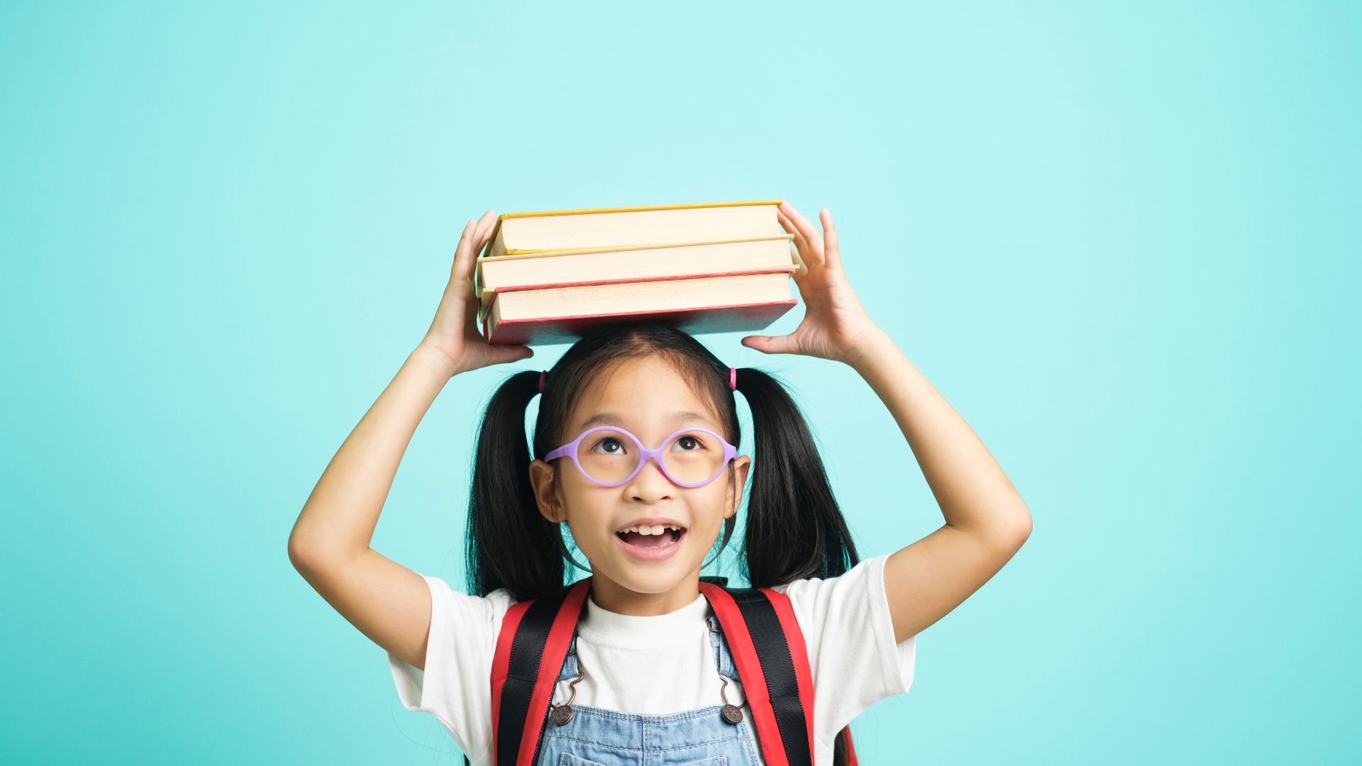 Ijeab-stock de iJeab grátis para o Canva Pro Imagem de uma criança estudante. Ela usa óculos de grau, mochila nas costas e equilibra três livros sobre a sua cabeça