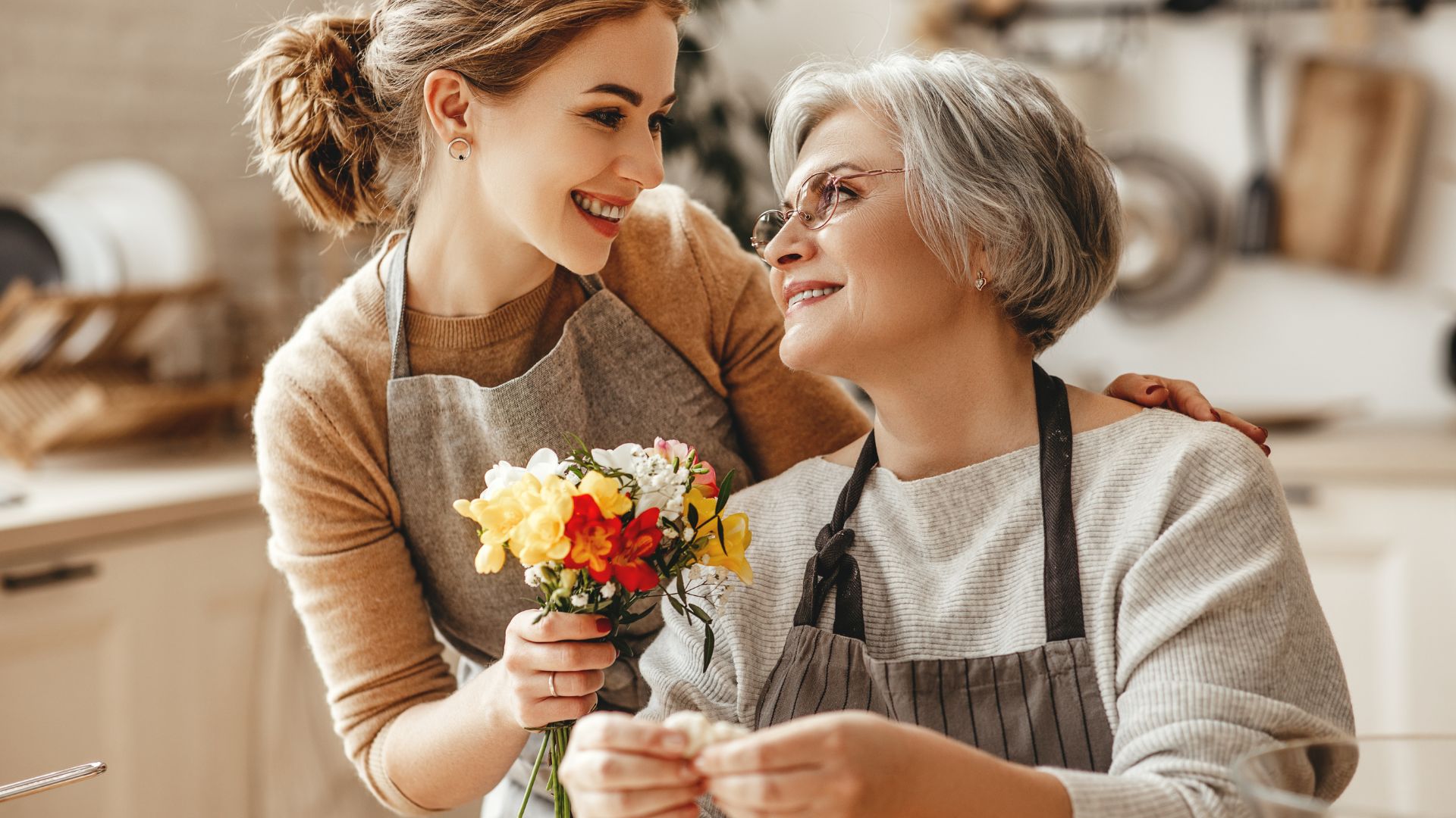 Evgenyatamanenko de Getty Images grátis para o Canva Pro Imagem de uma nora entregando flores para a sogra, demonstrando um gesto de carinho. Ambas estão felizes. Elas usam blusas de mangas longas nas cores bege e marrom e um avental.
