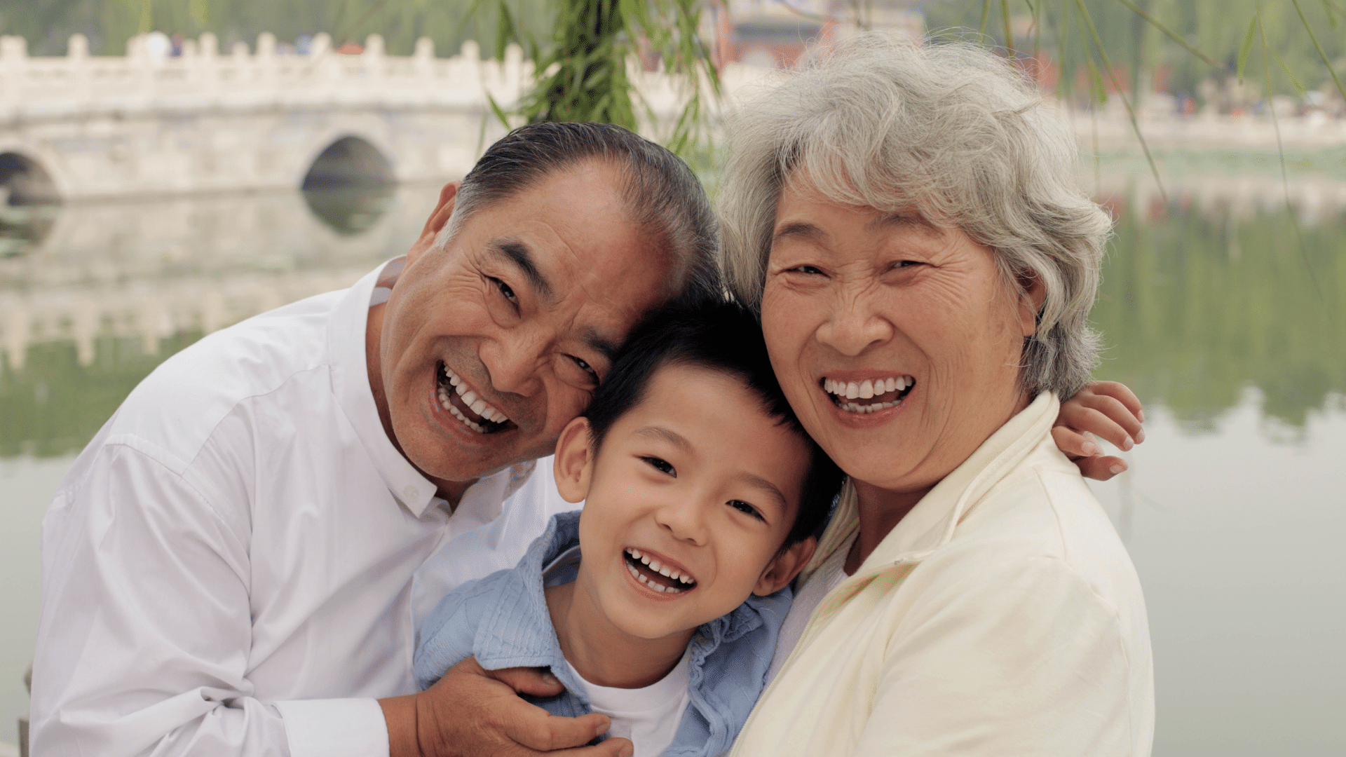 Imagens de avós abraçando seu neto na frente de um lago e uma ponte ao fundo da foto.