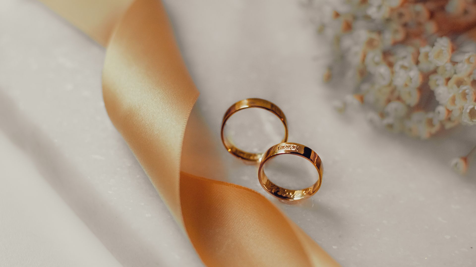 Imagem de duas alianças de casamento em cima de um tecido branco, ao lado de uma fita dourada e flores no tecido
