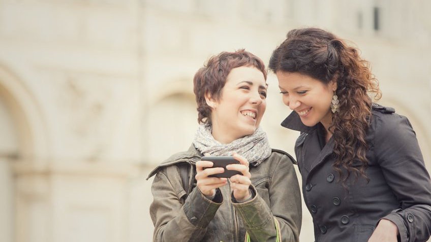 Duas mulheres sorrindo, e olhando para uma celular que uma delas segura.