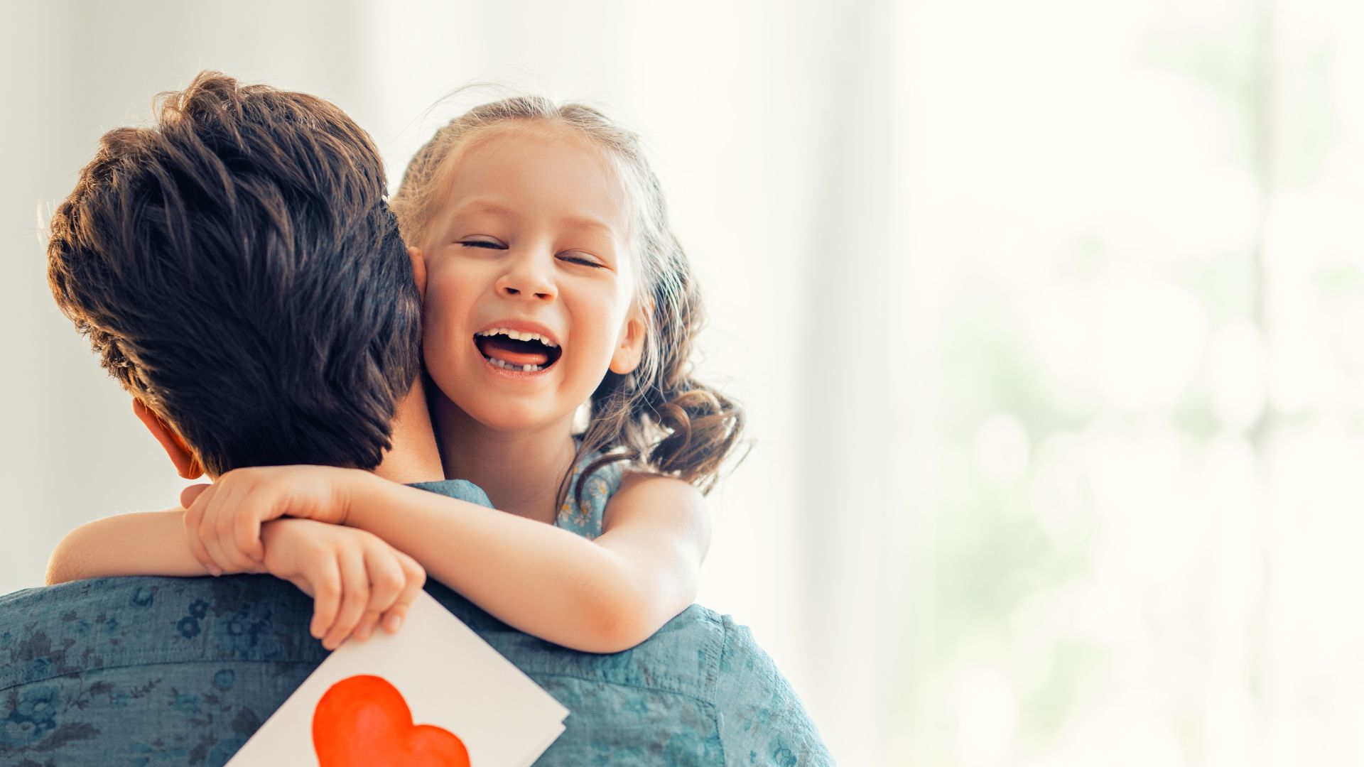 Imagem de um tio de costas, segurando sua sobrinha pequena no colo. Ela está abraçada no pescoço dele e segura em uma de suas mãos um cartão branco com um coração vermelho ao centro, desejando um feliz aniversário.